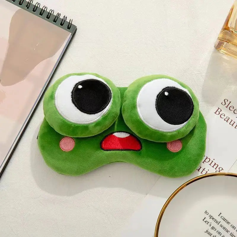 Green frog-shaped eye mask on a surface with a notebook and perfume bottle in the background.