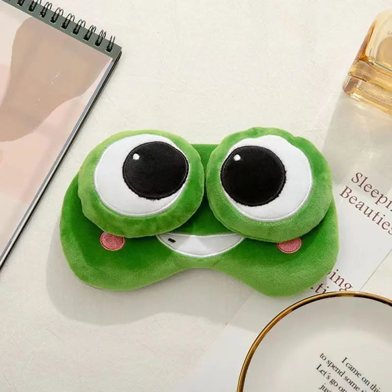 Green frog-shaped eye mask on a surface with a notebook and bottle in the background