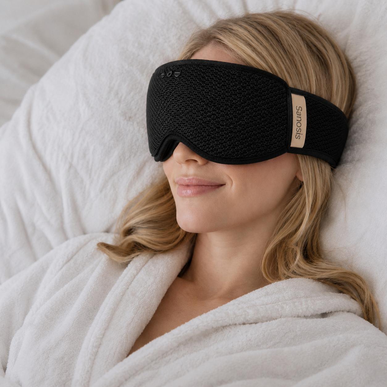 Woman wearing a black sleep mask with a brand logo, wrapped in white blanket.