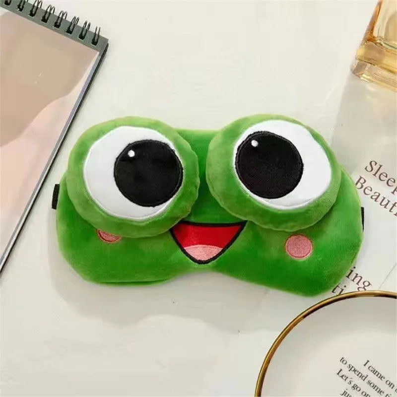 Green frog-shaped eye mask with large eyes and a smiling mouth on a surface.