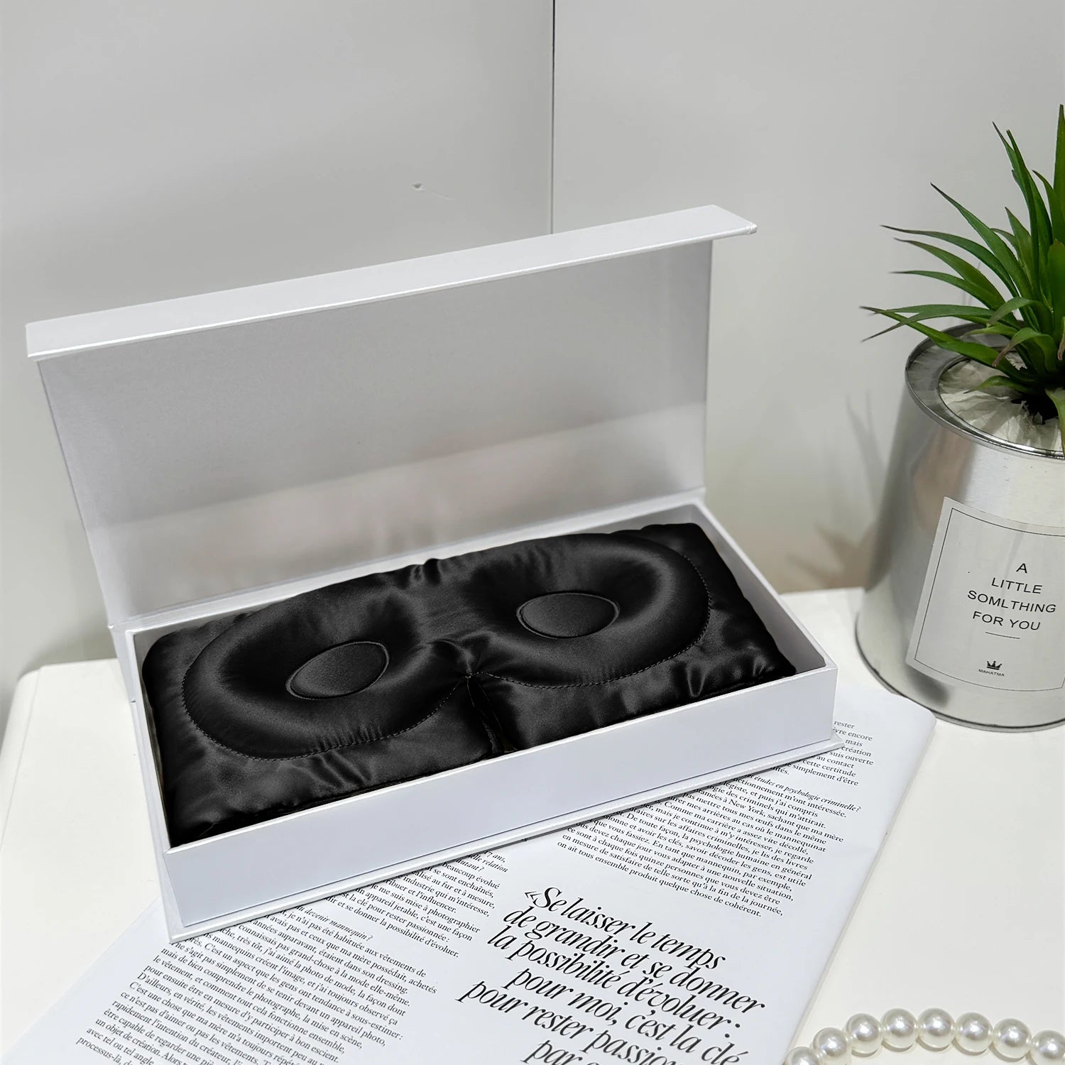 Black satin eye masks in a white box on a magazine with a plant and candle in the background.