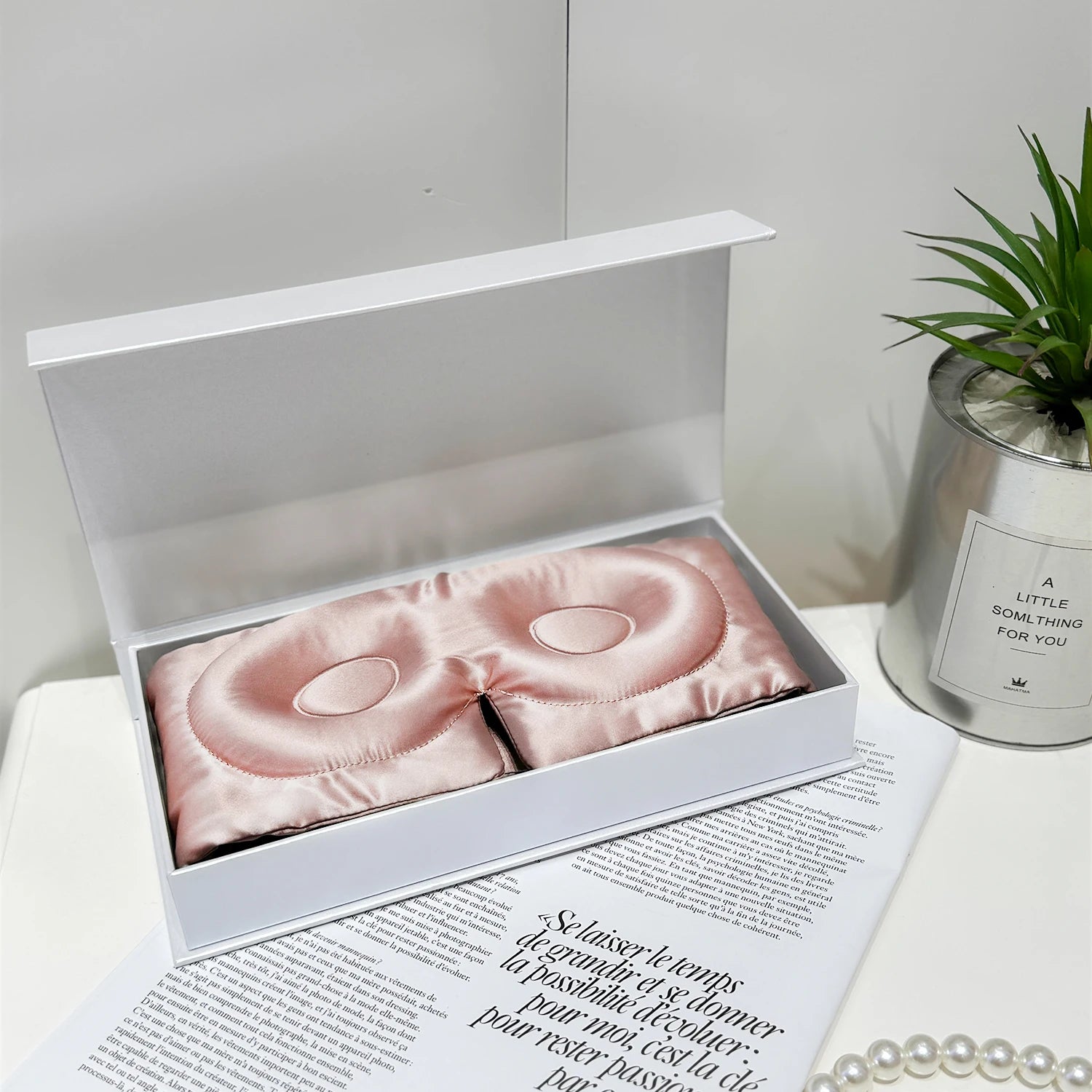 Pink satin eye mask in a white box on a magazine with a plant and candle in the background.